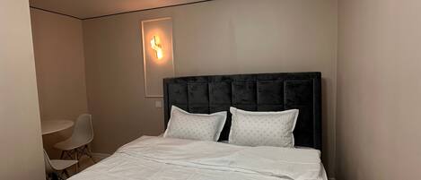 Een netjes opgemaakt bed met een donkere hoofdbord en twee kussens, een kleine tafel met een stoel, en een wandlamp.