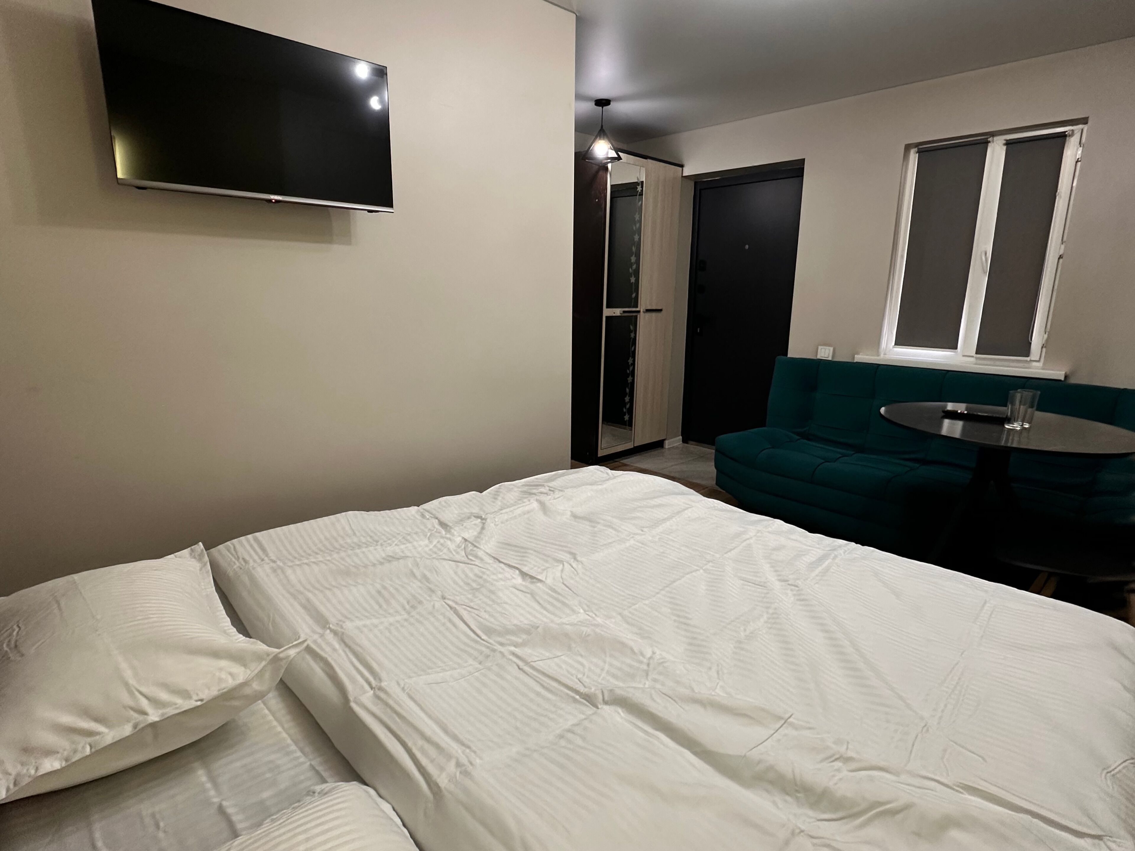 A bedroom with a bed, a TV mounted on the wall, a green sofa, and a small round table with a glass.