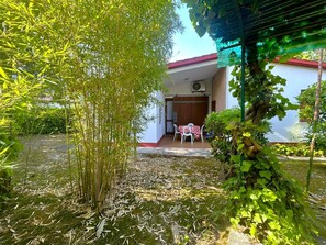A garden with a pond, a white building with a red roof, and a table with chairs outside. - DINKY VILLA 237 - B1 4+0 - C02372 (Bibione Pineda)