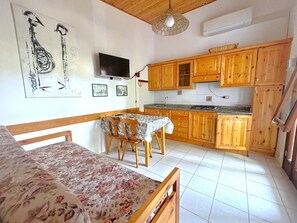 A compact kitchen with wooden cabinets, a dining table with chairs, a bed with a floral bedspread, and a mounted TV. - DINKY VILLA 237 - B1 4+0 - C02372 (Bibione Pineda)
