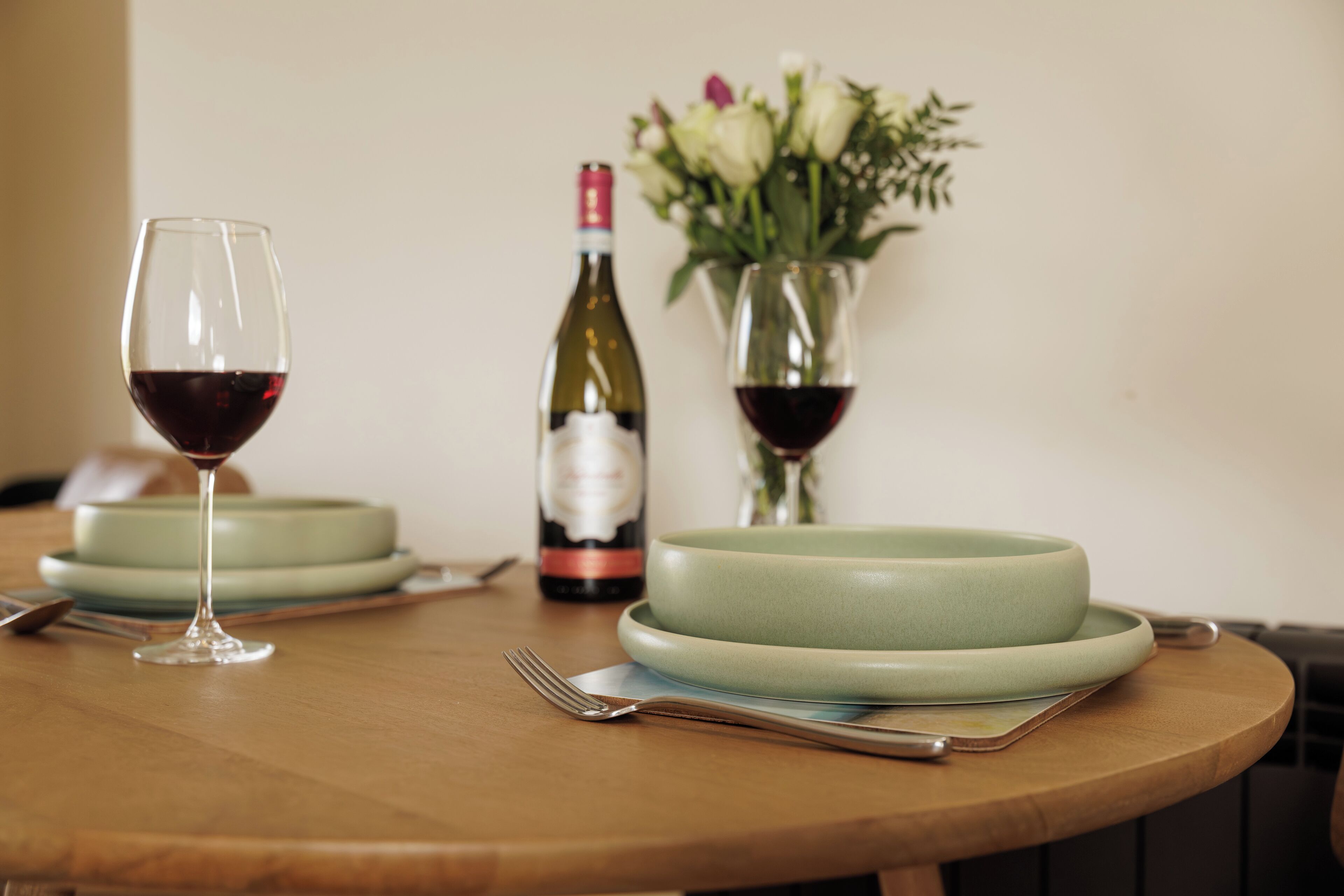 Una mesa de comedor con una botella de vino, dos copas de vino, un florero con flores y un plato.
