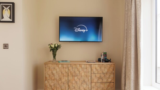 A television mounted on a wall displaying the Disney+ logo, a wooden cabinet with geometric patterns, a vase with flowers, and a window with curtains. -  Cosy Cottage with sea view, 10 mins from Portree (Portree)