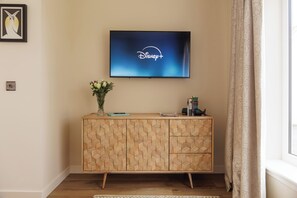 A television mounted on a wall displaying the Disney+ logo, a wooden cabinet with geometric patterns, a vase with flowers, and a window with curtains.