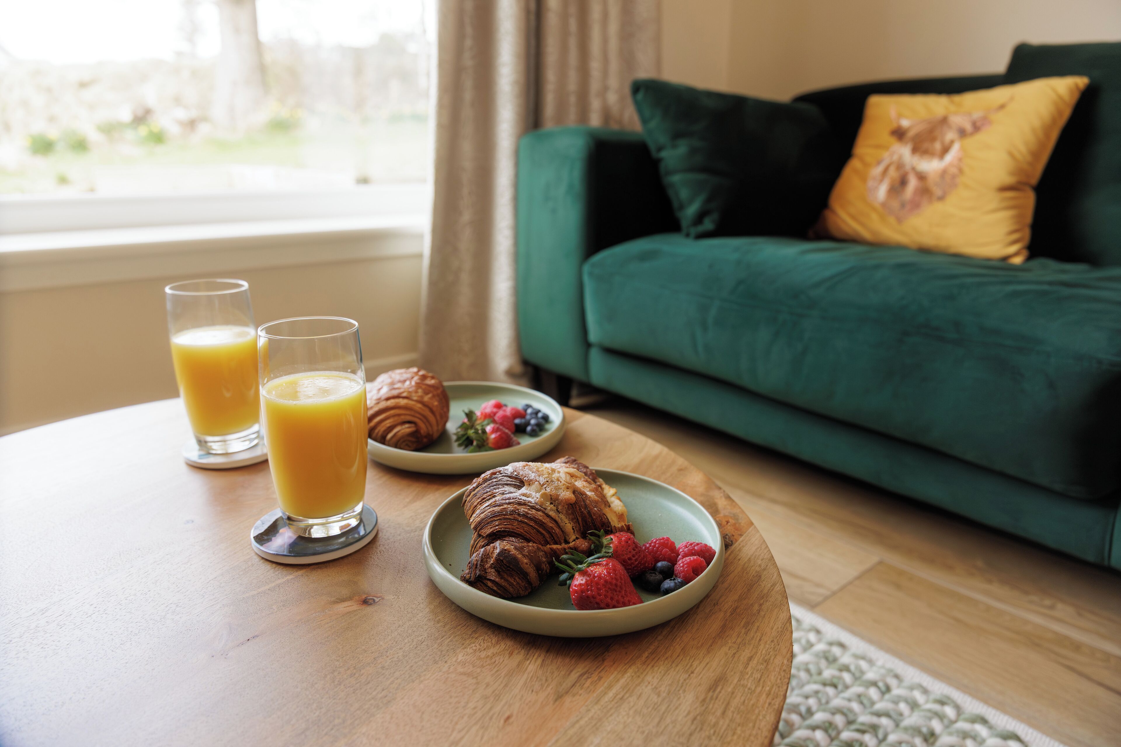 En frukostuppdukning med croissanter, bär och apelsinjuice på ett träbord, med en grön soffa och ett fönster i bakgrunden.