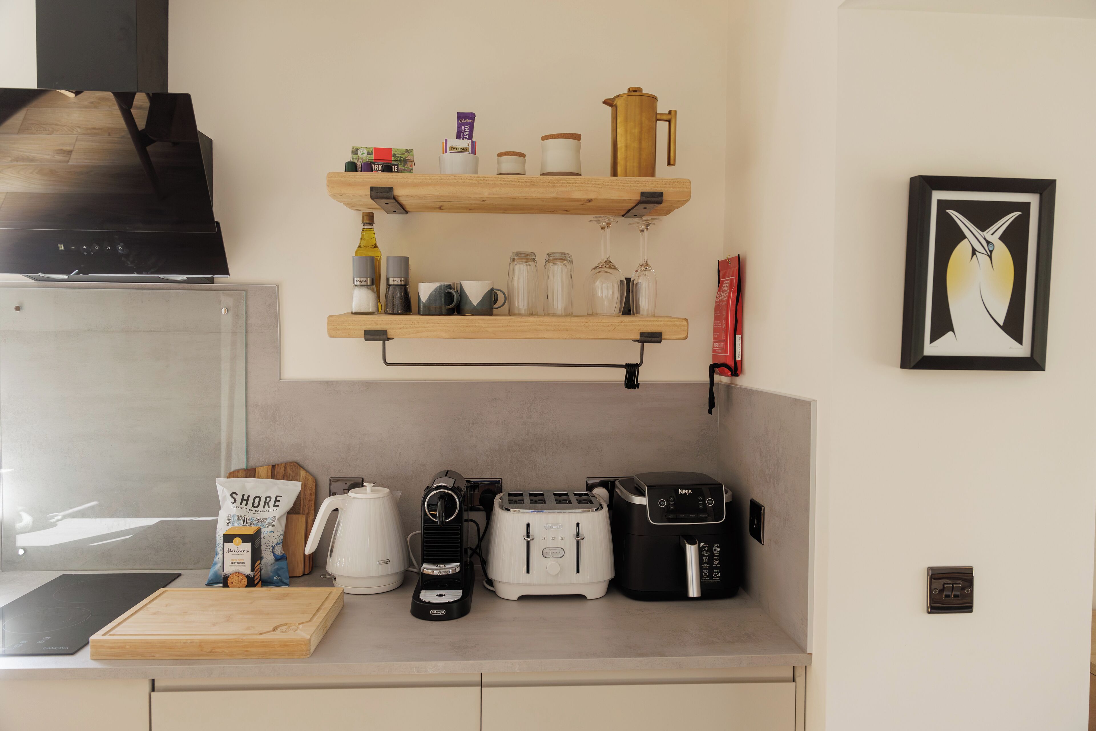 A kitchen with a coffee maker, toaster, and kettle on the counter, wooden shelves above with jars and a coffee pot, and a framed picture on the wall.