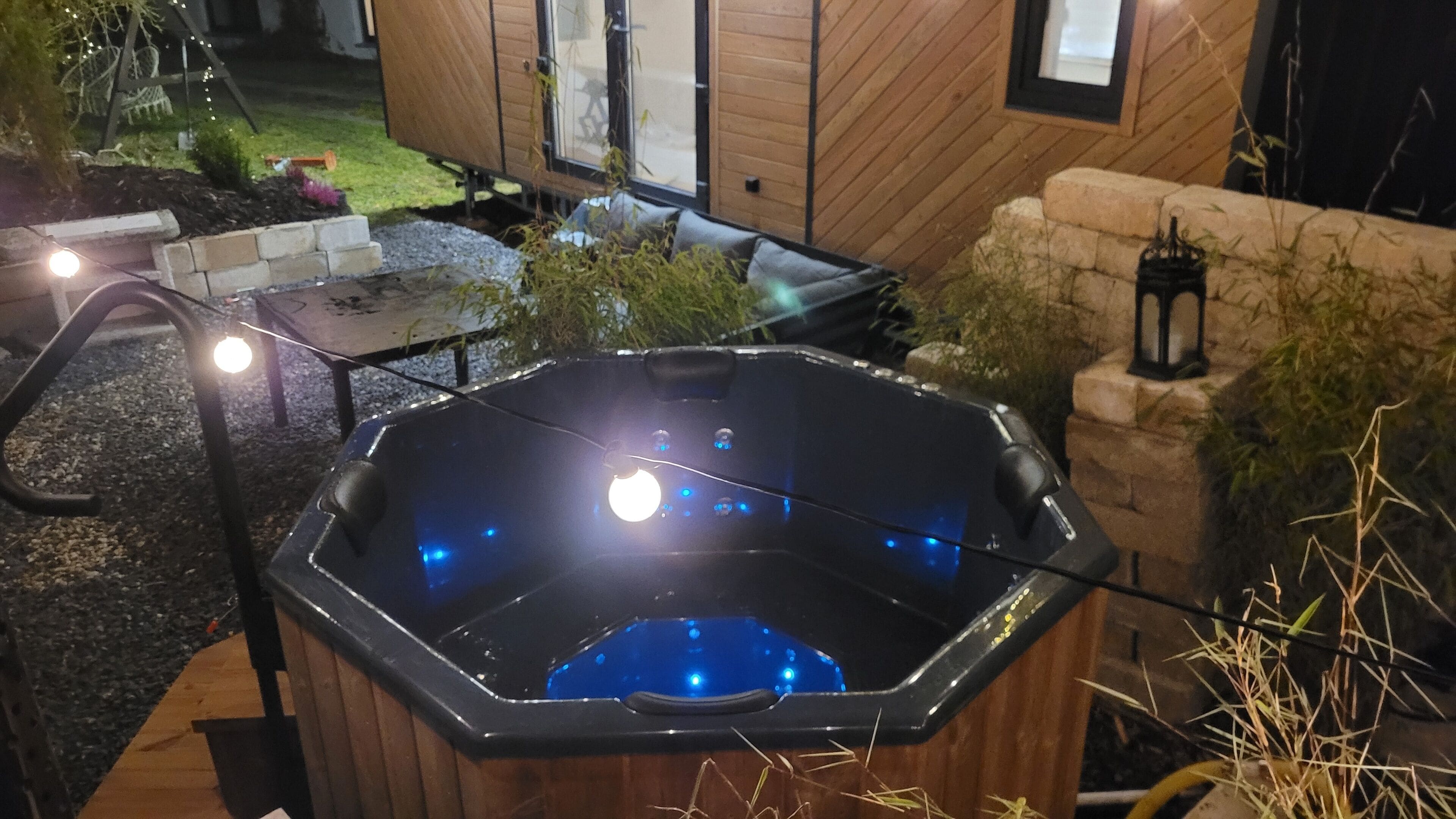 Ett bubbelbad med träpanel runt om, omgivet av en stenmur och en liten trädgårdsdel med växter och ett bord.