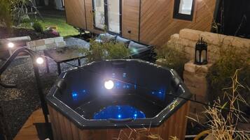 Ett bubbelbad med träpanel runt om, omgivet av en stenmur och en liten trädgårdsdel med växter och ett bord.