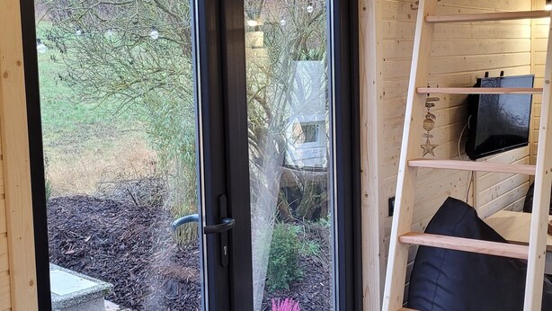 A cozy cabin interior with a sliding glass door leading to a garden, a wooden ladder, and a built-in TV shelf.