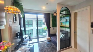 Una camera d'albergo moderna con un'ampia porta-finestra scorrevole che dà su un balcone, una zona pranzo con tavolo e sedie, e un angolo salotto accogliente con divano e lampada decorativa.
