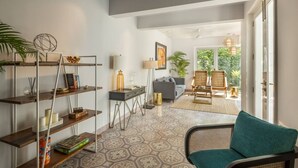Een ruime woonkamer met een bank, een salontafel en een boekenkast. Er is uitzicht op een tuin door de open deur.