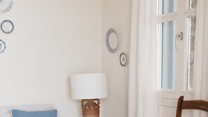 A room with a white sofa, a wooden rocking chair, a small table with a lamp, and a window with curtains. - Cycladic Cottage with sea access in a hideaway of rare character (Andros)