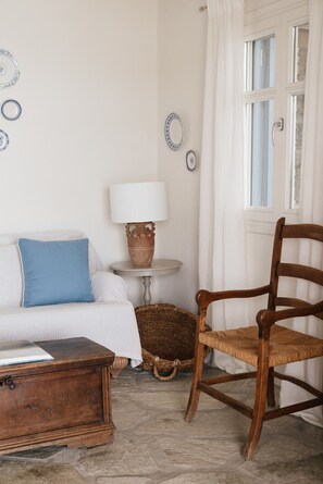 Ein Zimmer mit einem weißen Sofa, einem hölzernen Schaukelstuhl, einem kleinen Tisch mit Lampe und einem Fenster mit Vorhängen.