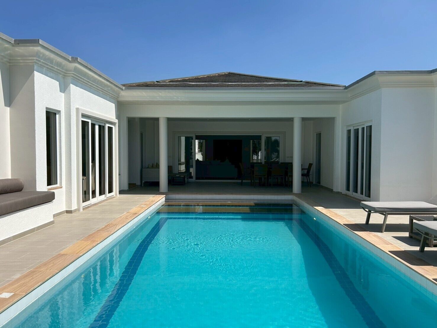 Piscina rectangular con solárium azulejado, rodeada por un porche cubierto con sillas y una mesa.