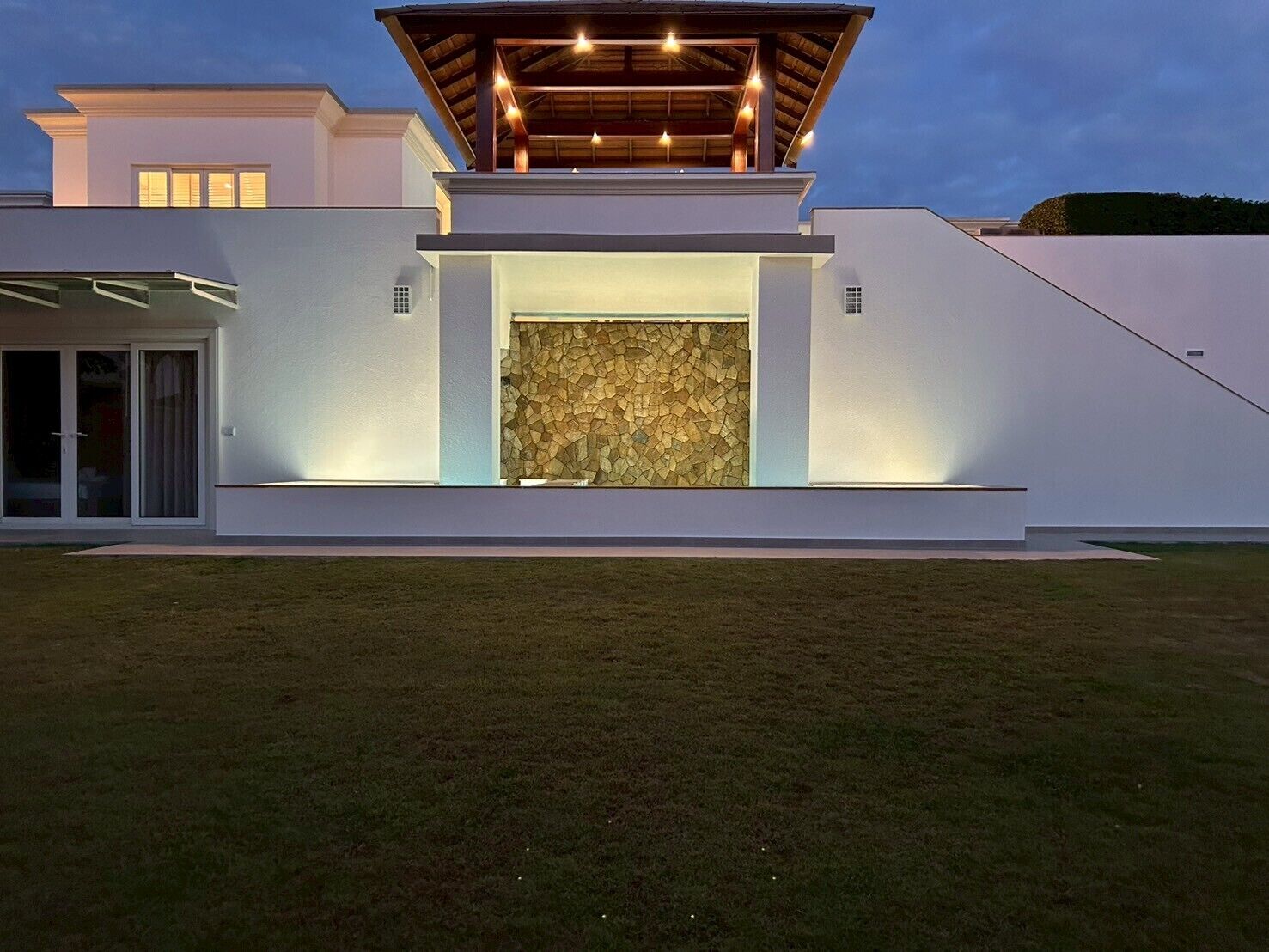 Una casa moderna con una imponente pared de piedra, un porche cubierto con luces y un césped bien cuidado.