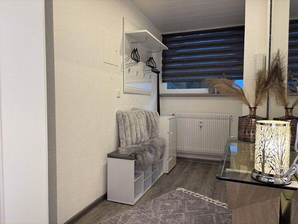 Habitació moderna i ben il·luminada amb un radiador blanc, una cadira mullada, una taula de vidre amb objectes decoratius i una finestra amb persianes.