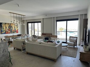 A modern living room with a large sofa, dining table, and chairs. There are large windows with a view of a pool and buildings. - Fanadir Marina 3-bedroom apartment El Gouna with an amazing Marina view (Red Sea Governorate)