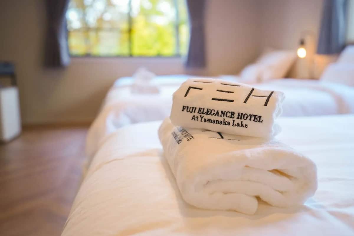 A neatly made bed with a towel folded on top, featuring the text "FUJI ELEGANCE HOTEL At Yamanaka Lake."