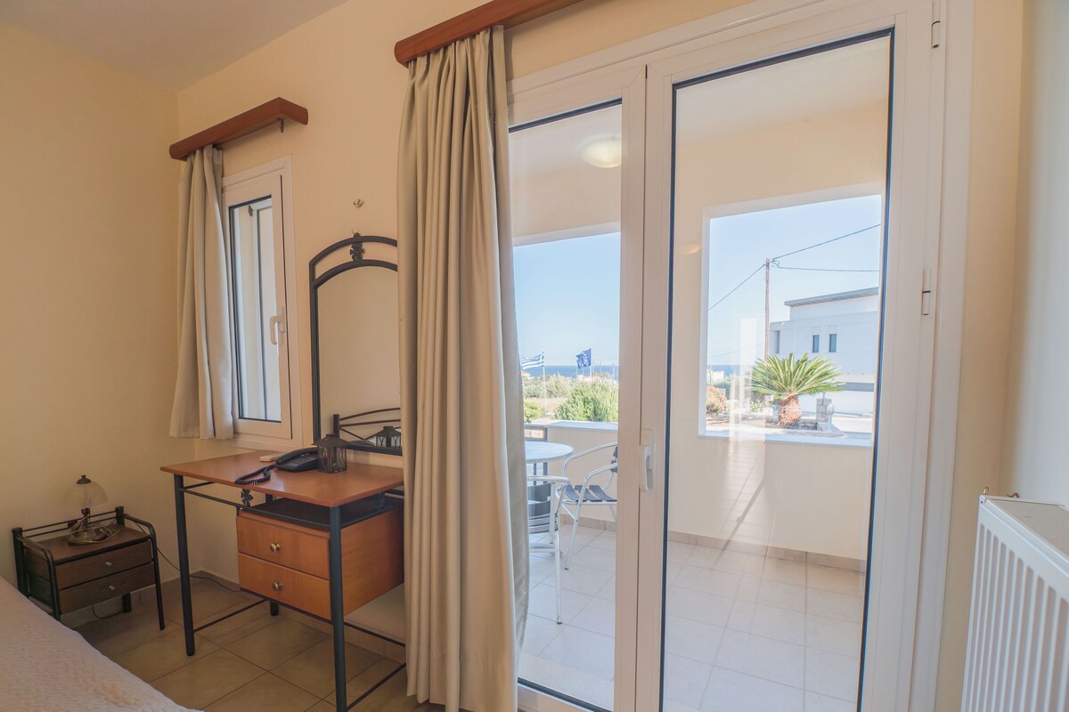 A room with a bed, a desk with a chair, a dresser, a mirror, and a sliding glass door leading to an outdoor balcony.