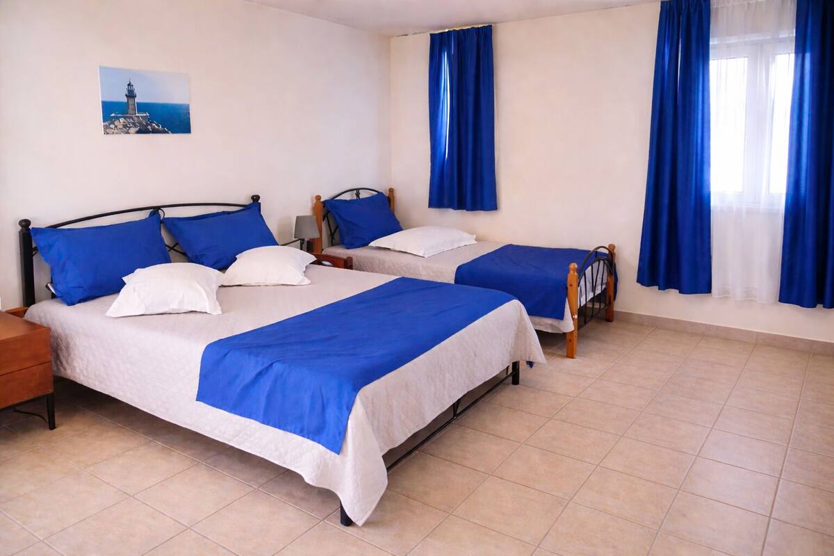 A hotel room with two beds, blue and white bedding, a nightstand, a lamp, and a painting of a lighthouse on the wall.