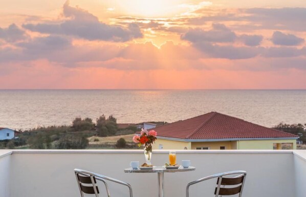 Un balcón con una mesa dispuesta para el desayuno, con vistas a un paisaje costero que incluye una casa y vegetación.