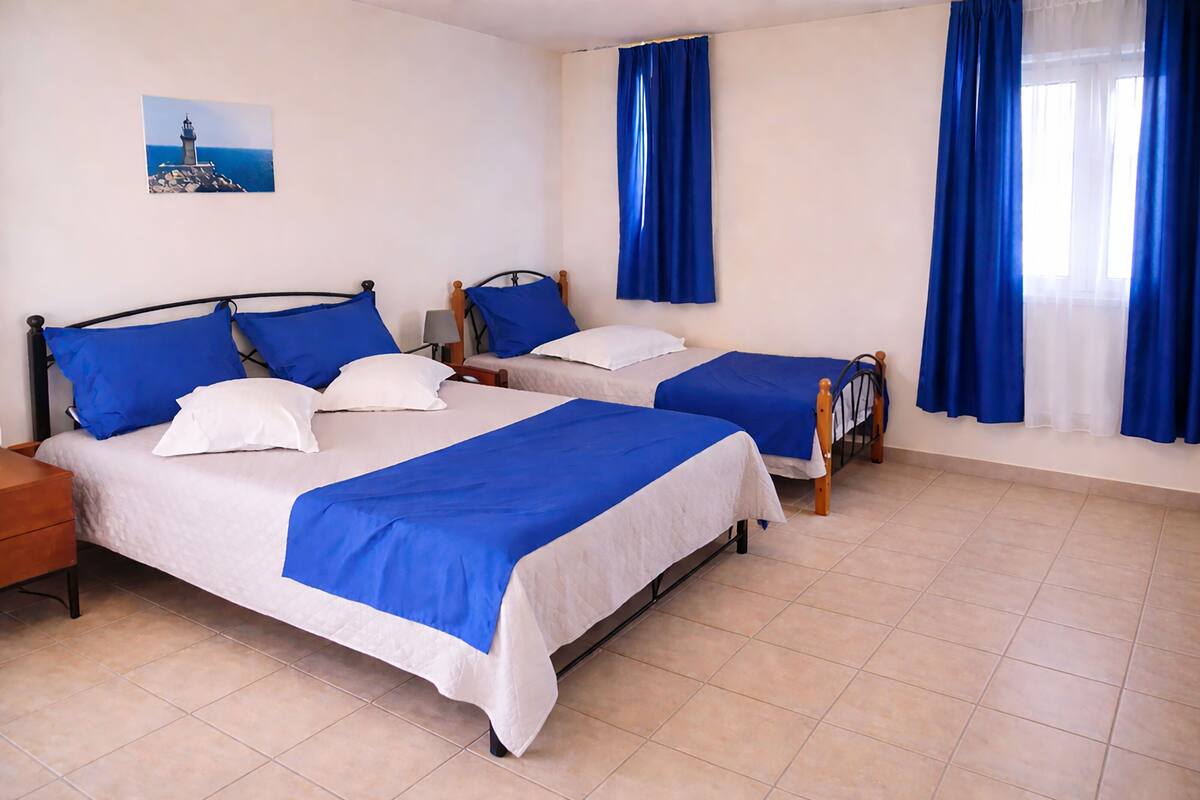 A room with two beds, each with a blue and white bedspread, a nightstand, a lamp, and a painting of a lighthouse on the wall.
