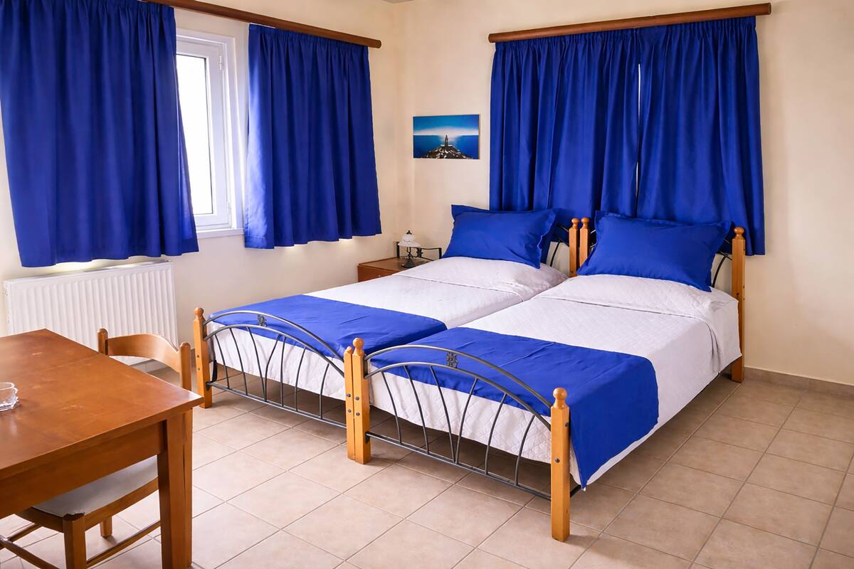 A double bed with a metal frame, a wooden table, and a chair in a room with blue curtains and a picture on the wall.