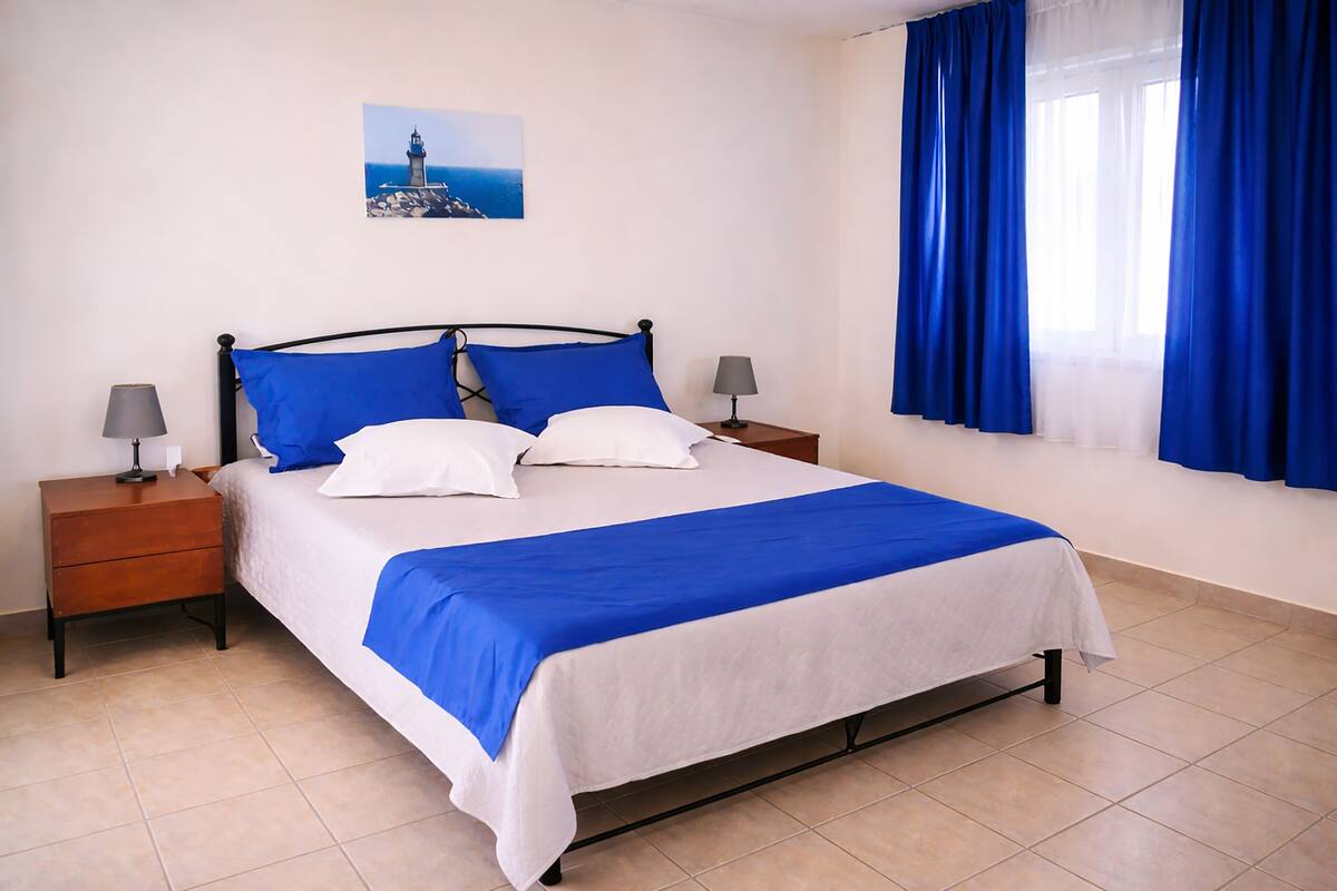 A bed with white and blue bedding, two bedside tables with lamps, a picture on the wall, and a window with blue curtains.