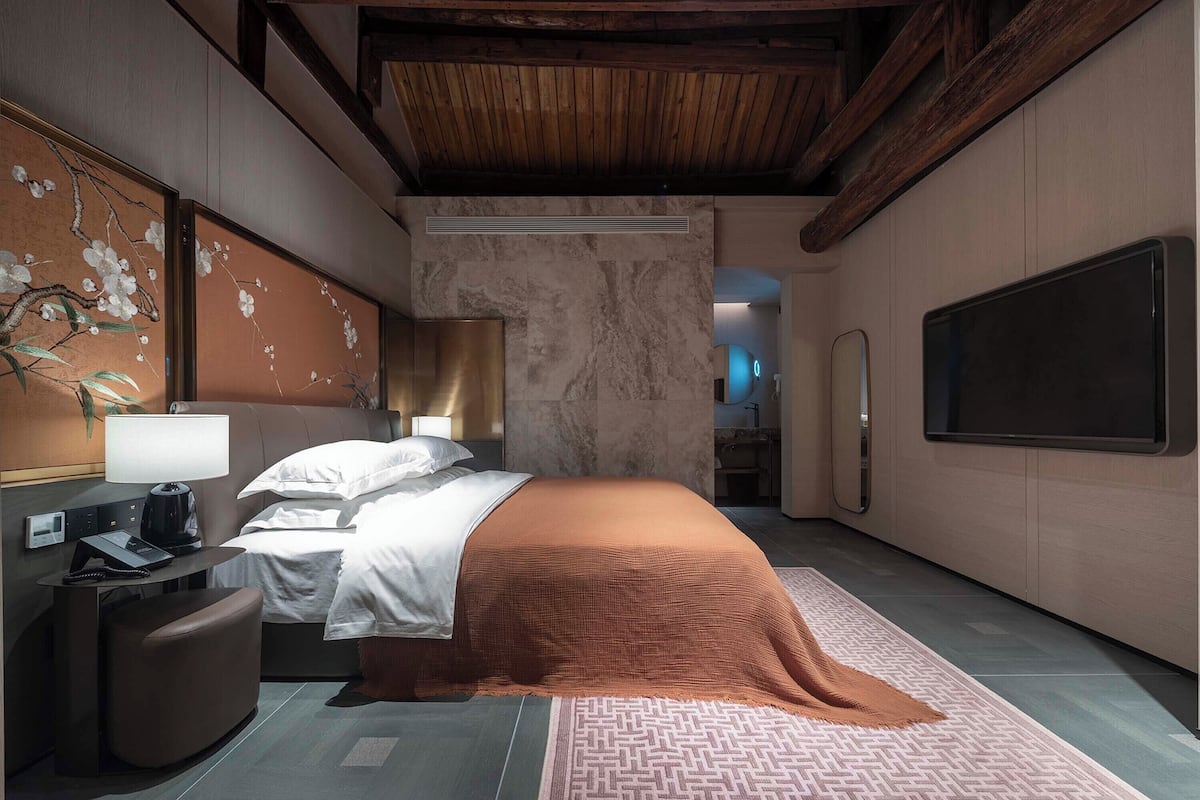 Una habitación de hotel moderna con una cama, un escritorio, una silla, un televisor y un baño visible a través de una puerta abierta.