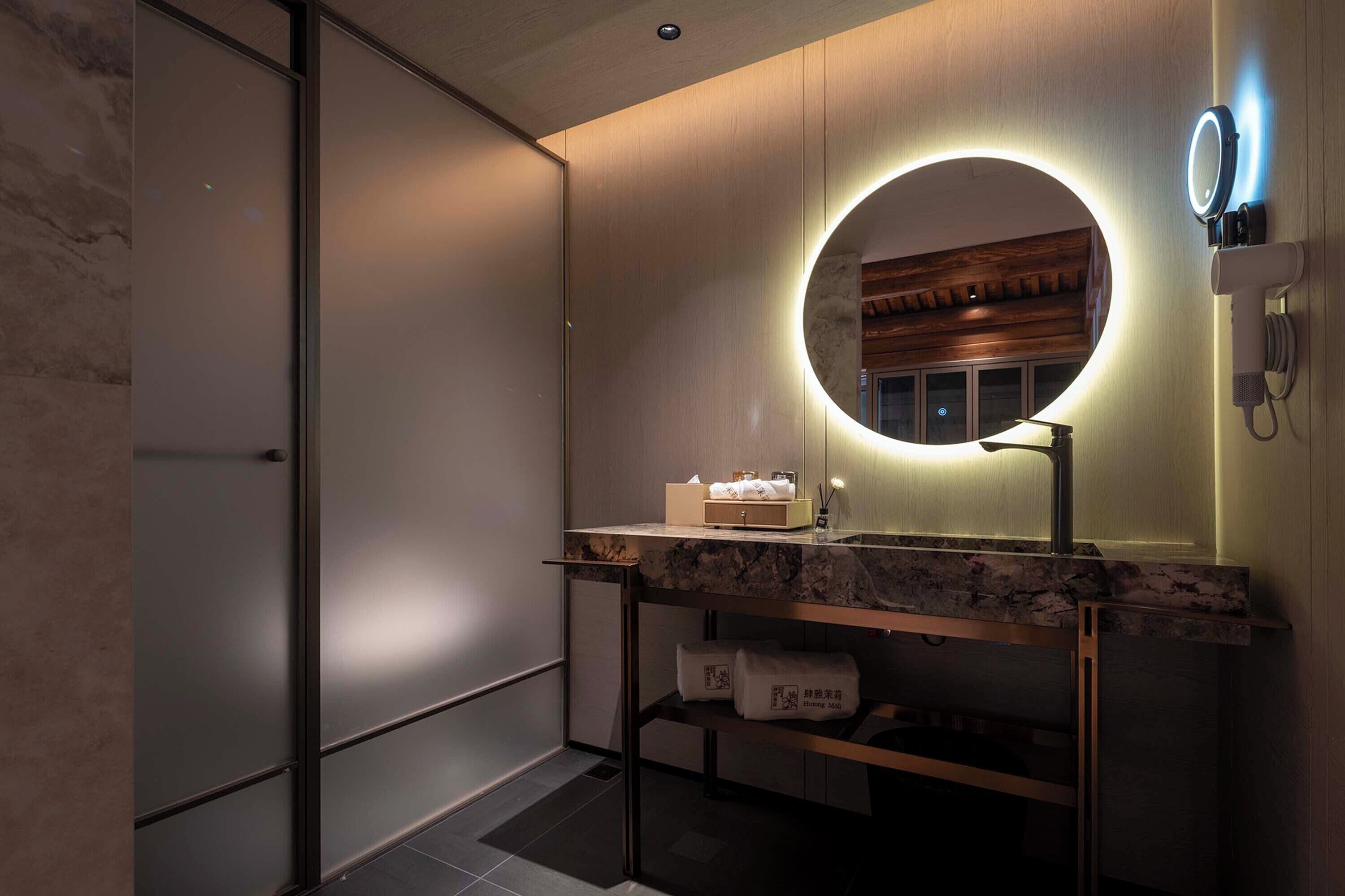 A modern bathroom with a marble countertop, a round mirror with a light, and a wooden vanity.