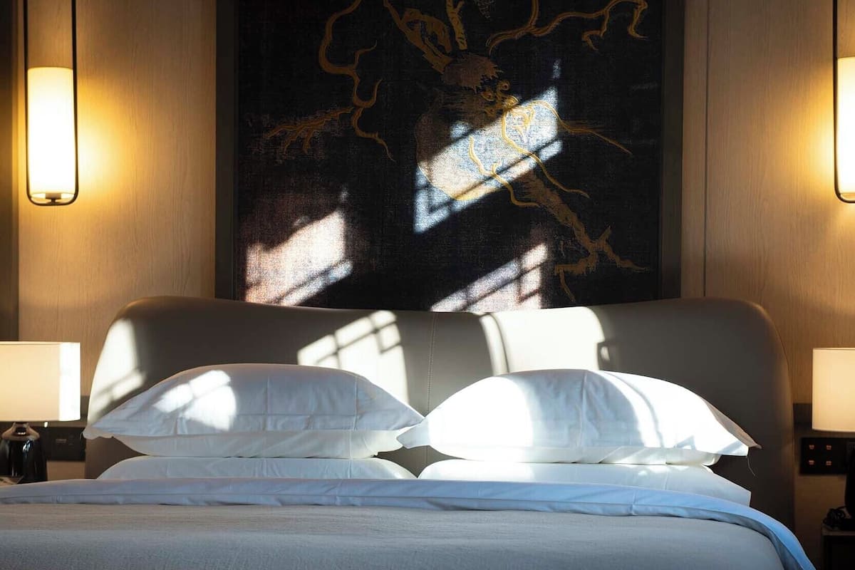 Una cama bien hecha con sábanas y almohadas blancas, un cuadro abstracto grande en la pared y dos lámparas de pared a cada lado.