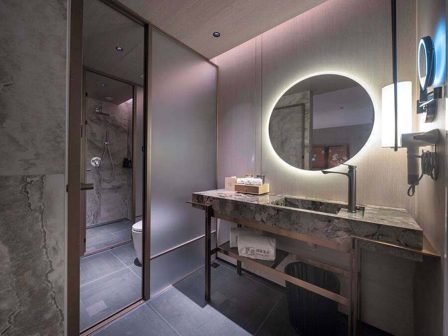 A modern bathroom with a marble vanity, a round mirror with integrated lighting, a handheld showerhead, and a towel rack.