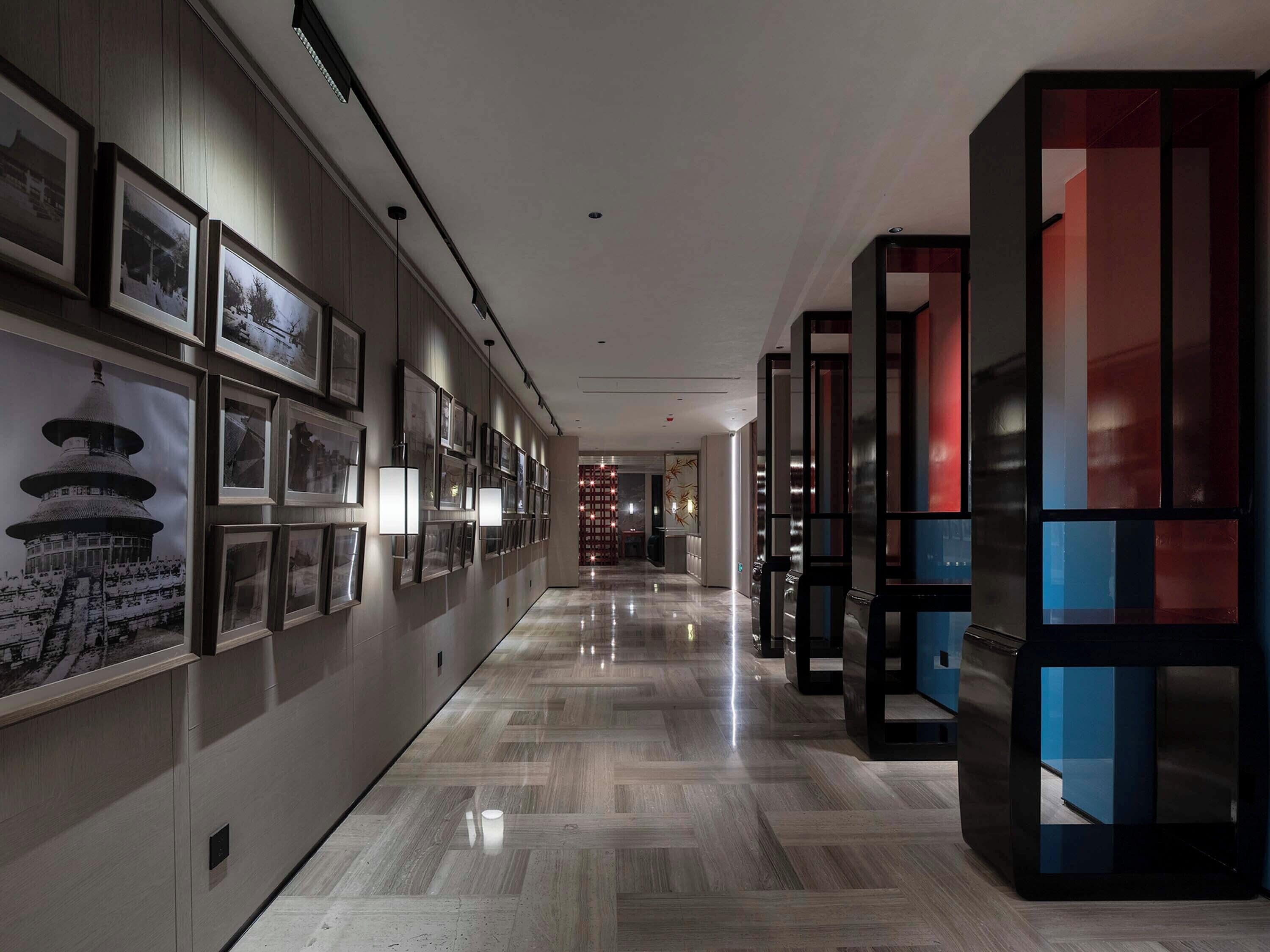 A hallway with framed pictures on the left, a modern elevator on the right, and a polished marble floor.