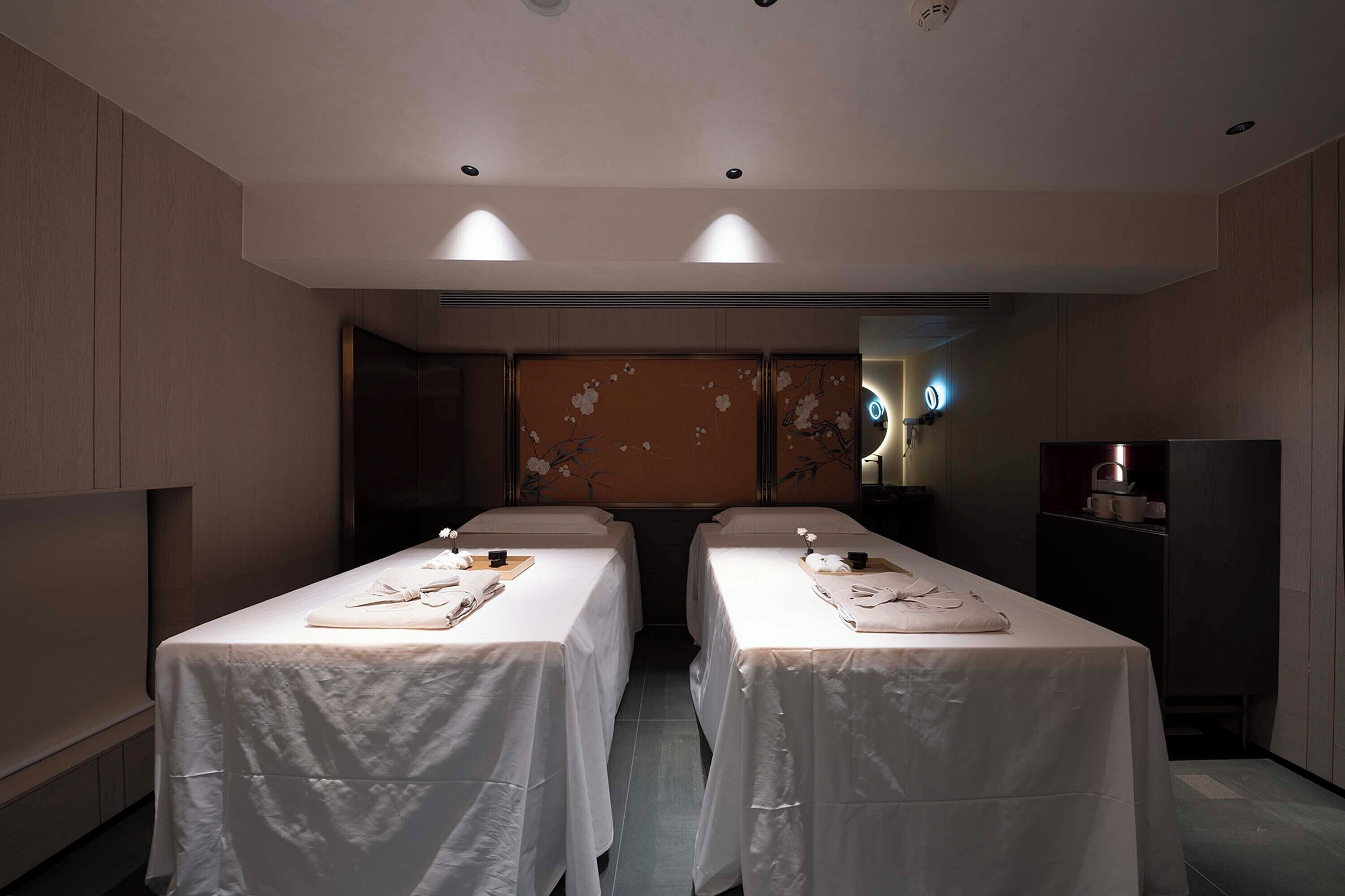 A hotel room with two beds, each with a white bedspread and a small table with a pillow and a cup. The room has a wooden panel wall and a decorative panel with floral designs.