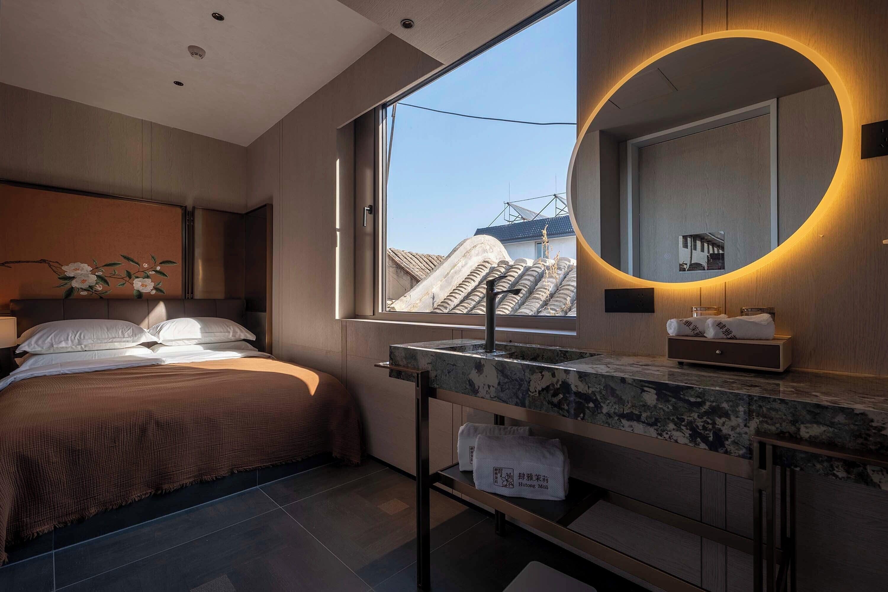 Ein modernes Hotelzimmer mit einem großen Bett, einem runden Spiegel mit warmem Licht, einem Badezimmer mit Marmorplatte und Blick auf Gebäude vor dem Fenster.