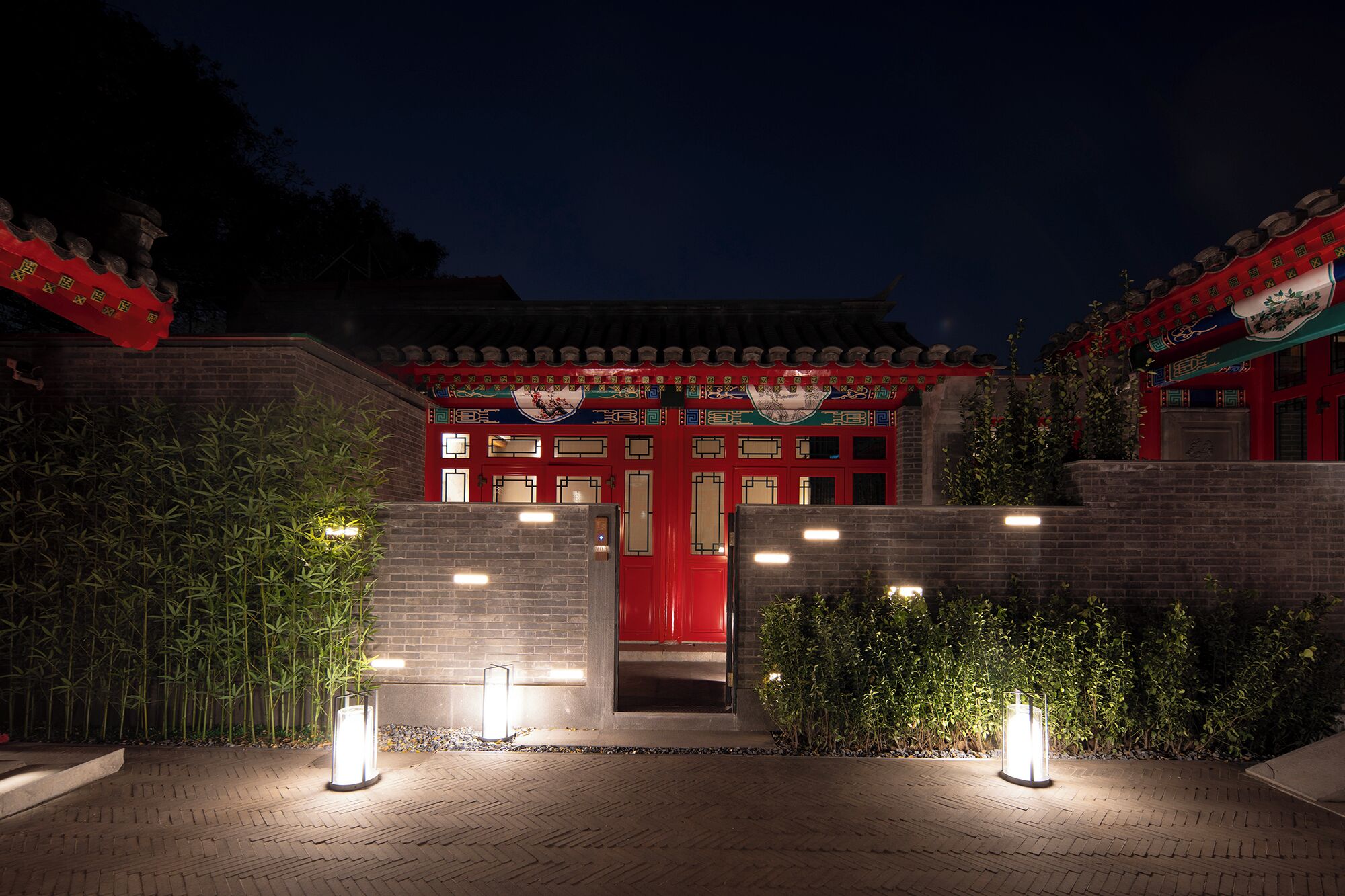 A traditional building with a red entrance, illuminated by ground lights, surrounded by bamboo and shrubs.
