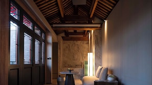 A hotel room with a bed, a small round table with a fruit bowl, a nightstand with a lamp, and a large window with red blinds. - Hutong Mòlì Hotel (Beijing)
