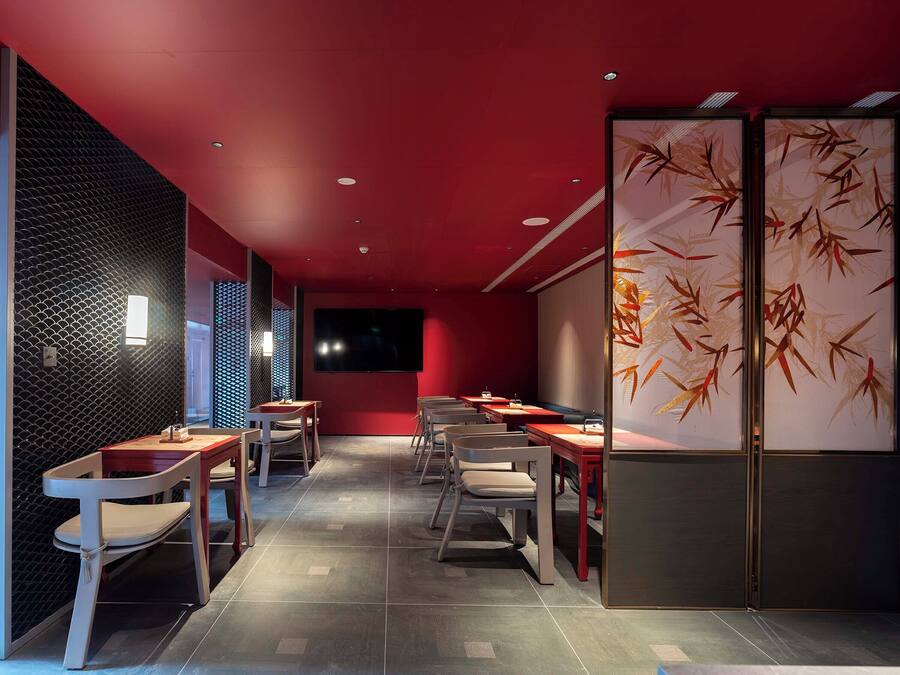 Ein modernes Restaurant mit roten Wänden, Wandverkleidungen mit schwarzem Muster und einem großen Bildschirm. Tische und Stühle sind zum Essen angeordnet.