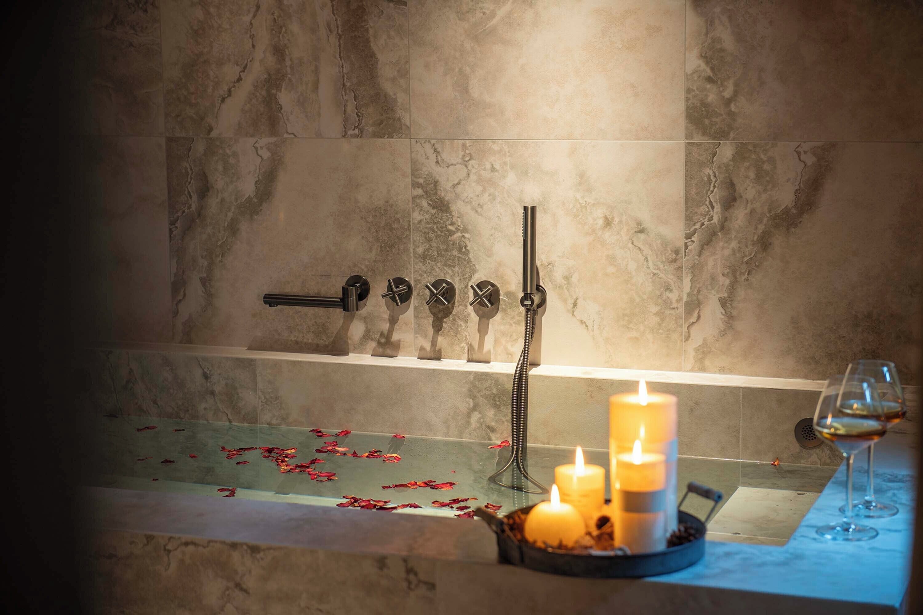 A bathroom with a bathtub, a set of showerheads, candles, and a glass of wine on the counter.