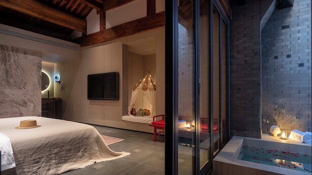 A modern bedroom with a large bed, a glass-enclosed window, a small balcony with a red chair, and a bathtub with rose petals. - Hutong Mòlì Hotel (Beijing)