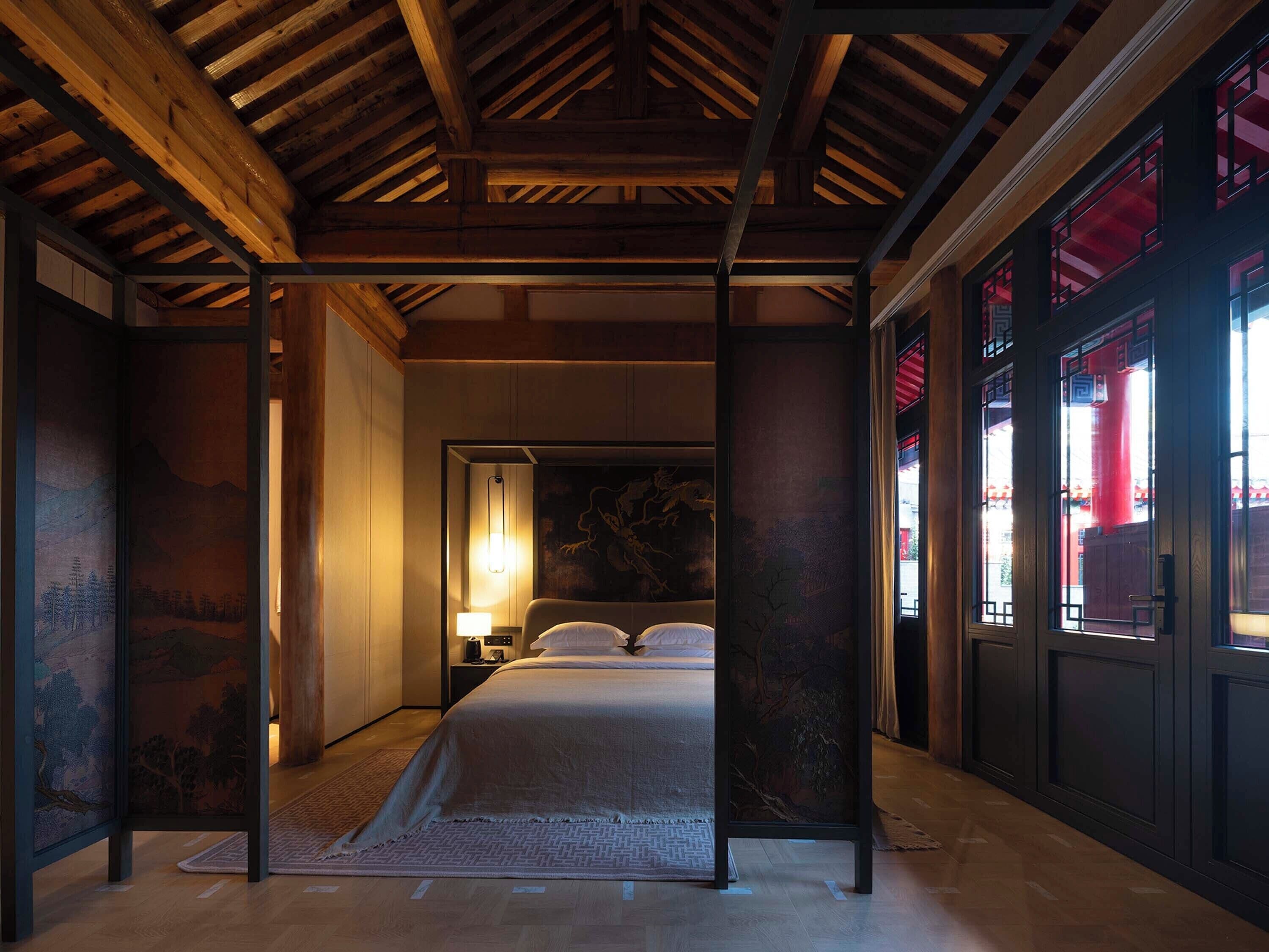 A traditional-style bedroom with a wooden ceiling, a bed with a white bedspread, a nightstand with a lamp, and a view of the outside through a window with red curtains.