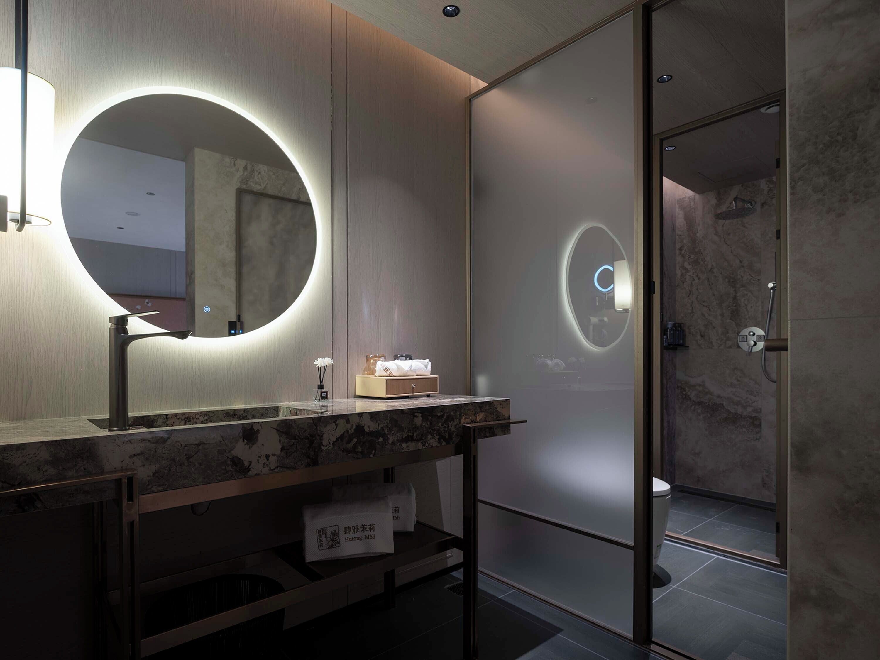 A modern bathroom with a marble vanity, a round mirror with integrated lighting, a glass shower enclosure, and a toilet.