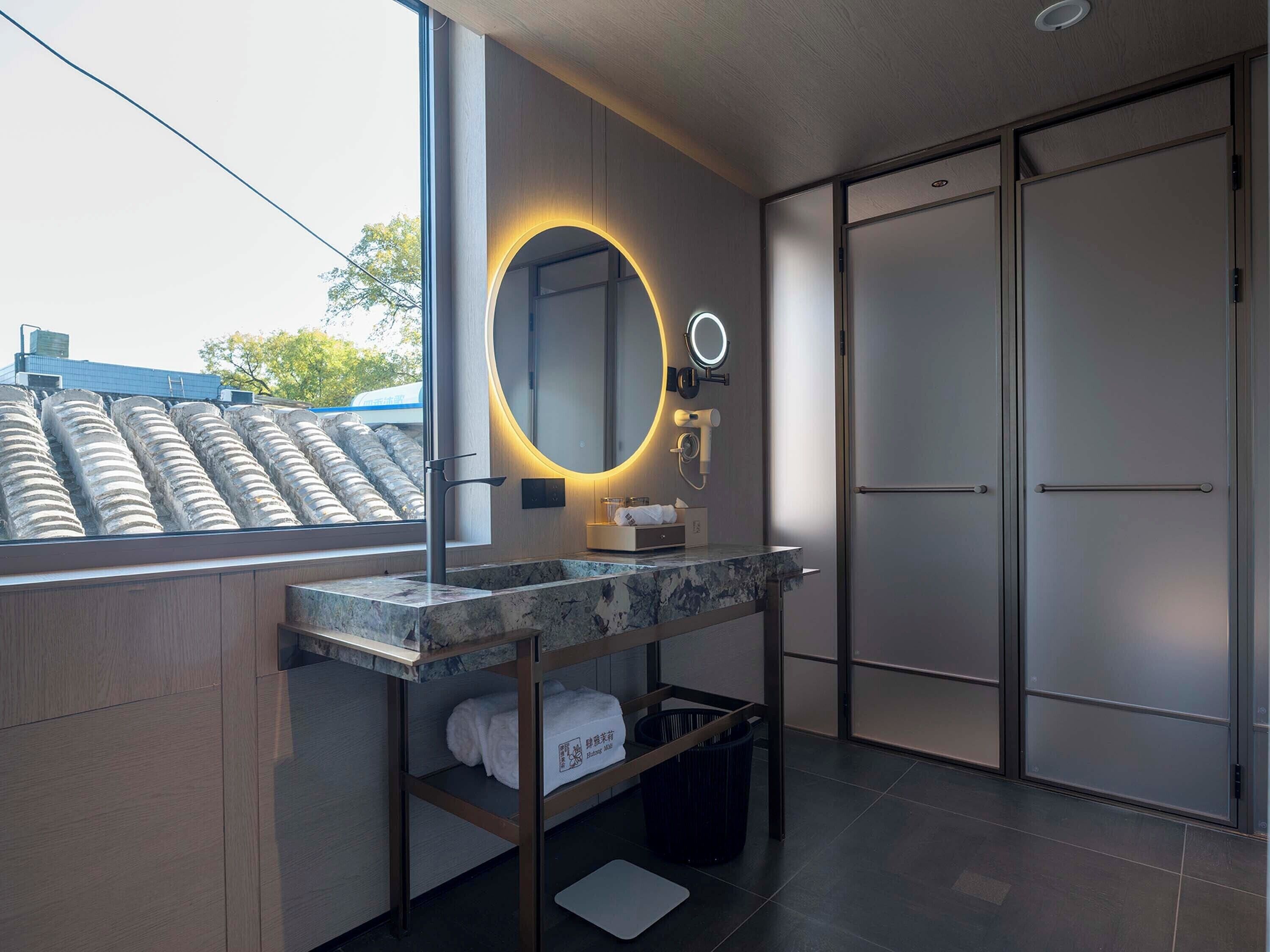 A modern bathroom with a large mirror, a vanity with a marble countertop, a towel rack, and a view of the outside.