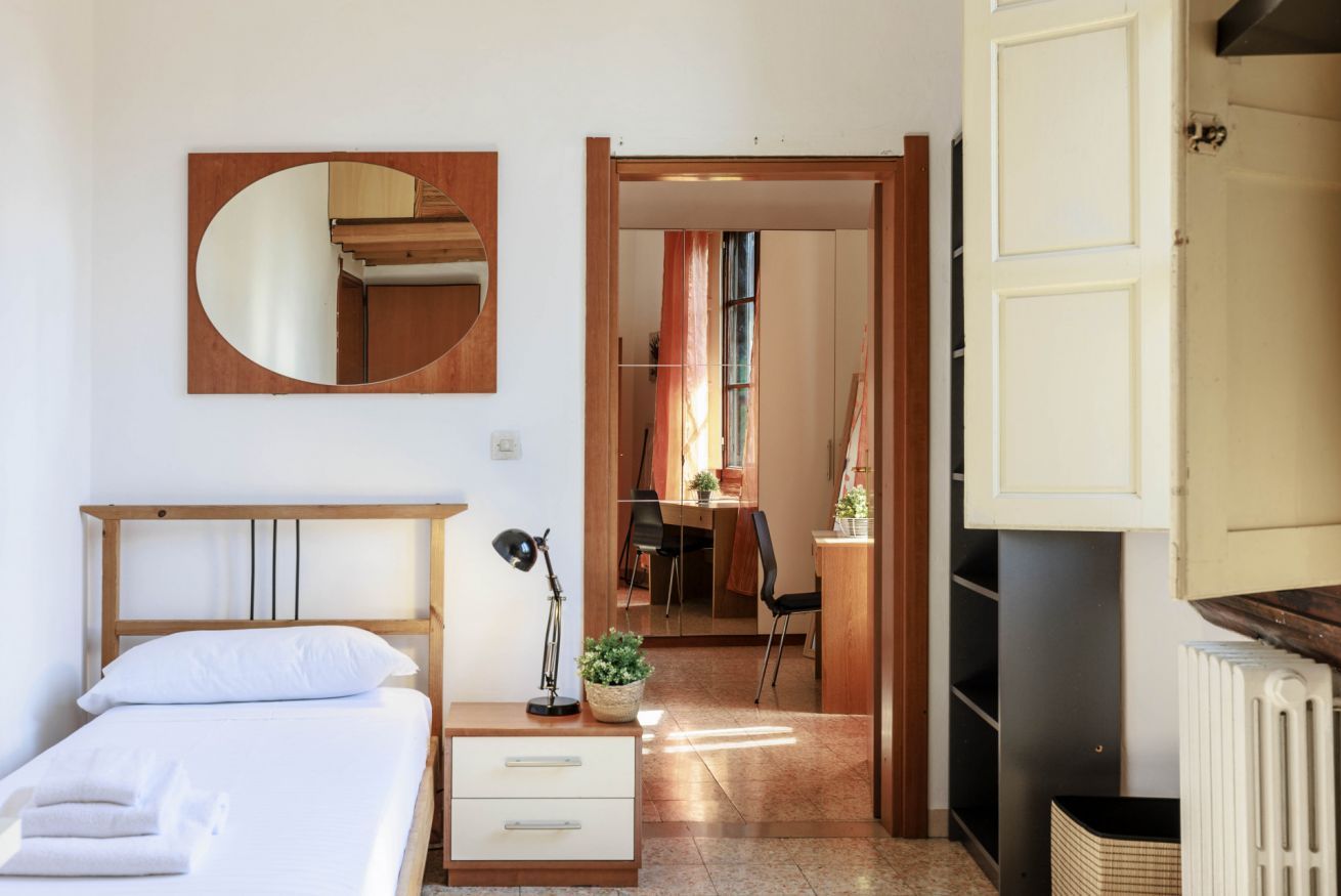 A hotel room with a bed, bedside table, lamp, and a mirror. There is a door leading to a dining area with chairs and a table.