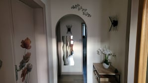 A hallway with a wooden floor, a door with a floral decoration, a mirror with a wavy frame, and a small table with a vase of flowers. - Bright ground-floor apartment with garden. (Gérardmer)