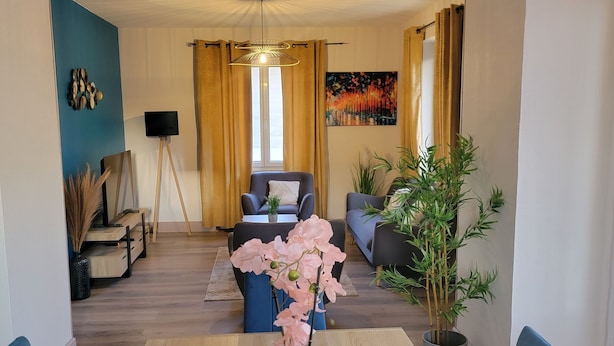 A dining area with a wooden table, blue chairs, and a vase of pink flowers. There is a sofa and a coffee table in the background, along with a potted plant and a painting on the wall. - Bright ground-floor apartment with garden. (Gérardmer)