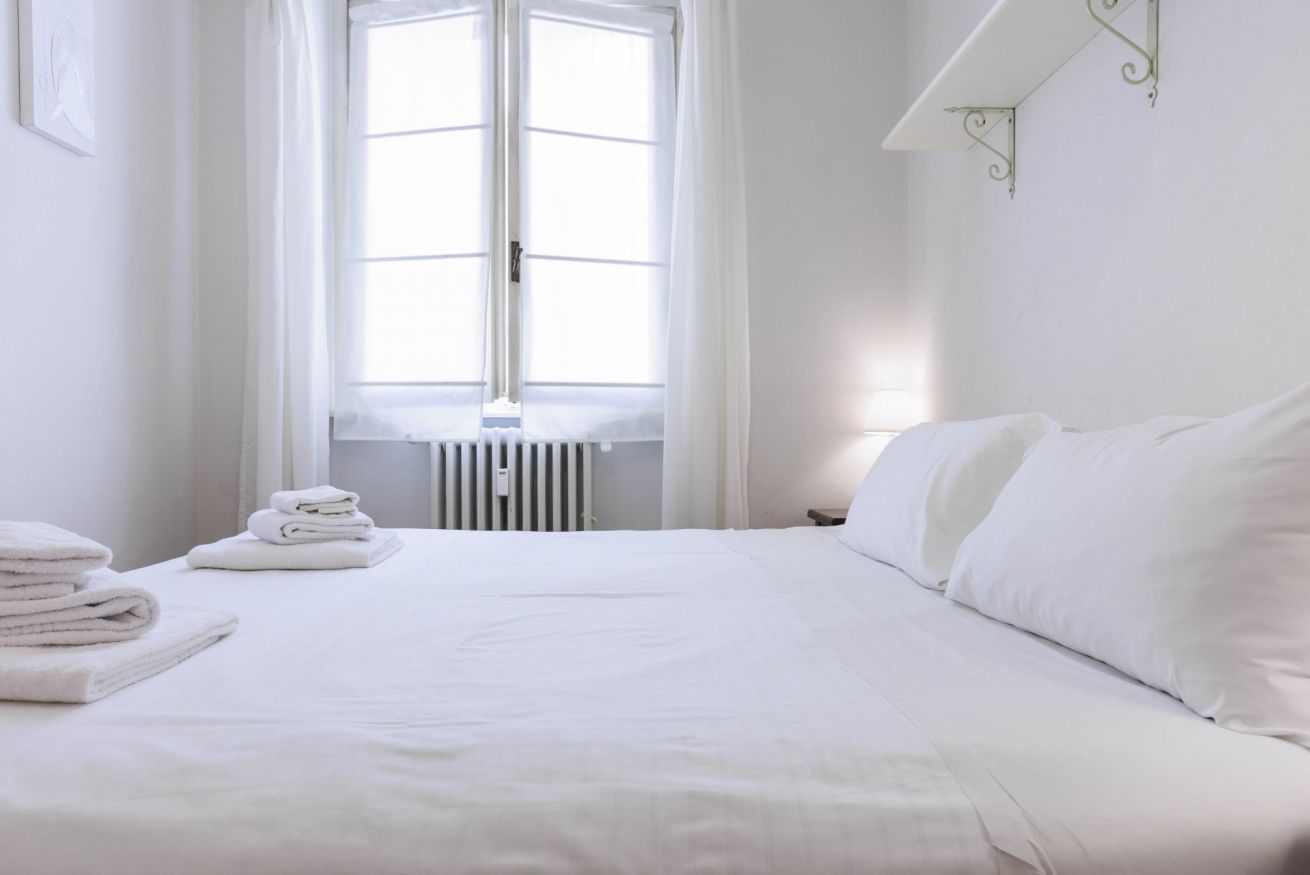 A neatly made bed with white linens and a radiator below a window.
