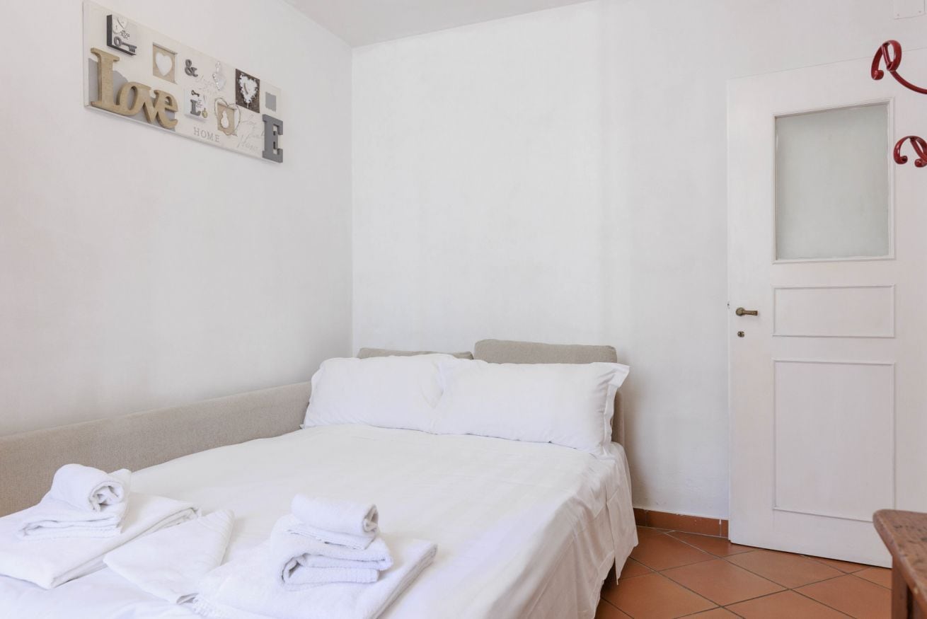 Uma cama bem arrumada com roupas de cama e toalhas brancas, uma porta branca com um pequeno espelho e um enfeite de parede com a palavra “Love” e outras letras.