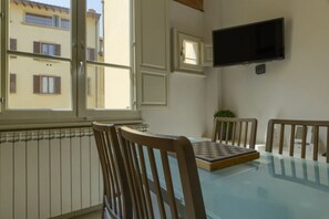 Ein Essbereich mit einem Glastisch, Holzstühlen, einem Schachbrett, einem an der Wand befestigten Fernseher und einem Fenster mit Blick auf Gebäude.