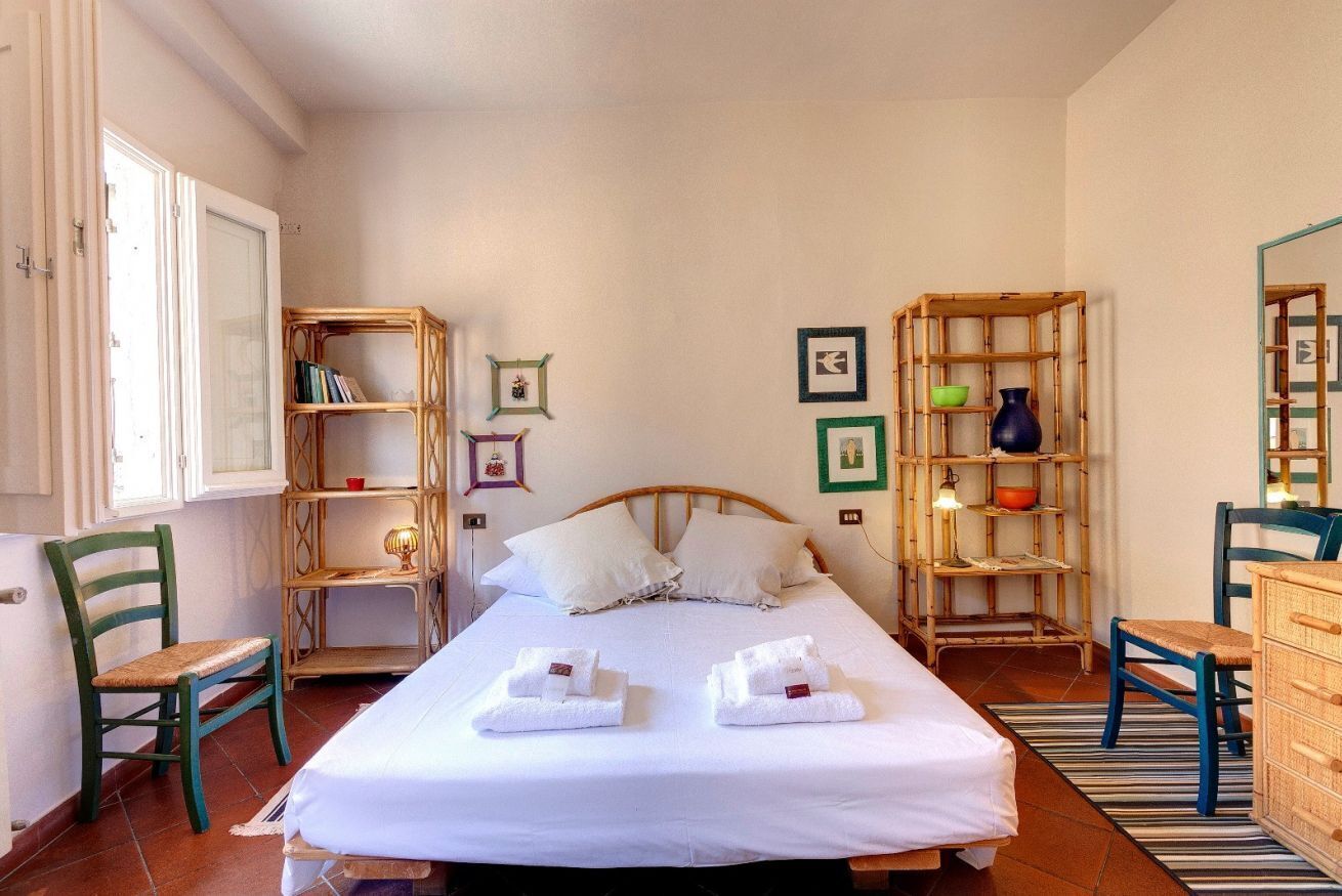 Un dormitorio ordenado con una cama, dos sillones, un aparador y dos estantes con libros y objetos decorativos.