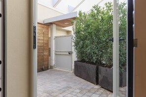 Um corredor exterior estreito com chão em azulejos, uma porta de madeira e dois grandes vasos com folhagem verde.